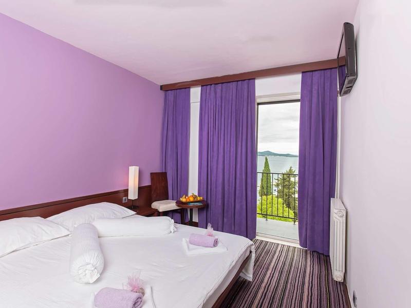 Moderní hotelový pokoj s manželskou postelí, fialovými stěnami a výhledem na balkon a krajinu.