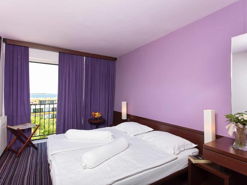 Světle fialový hotelový pokoj s manželskou postelí, balkonem a moderním zařízením.