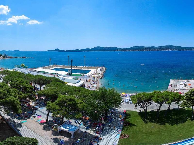 Panoramatický výhled na pobřeží s modrým mořem, zeleným parkem a mnoha loděmi v přístavu.