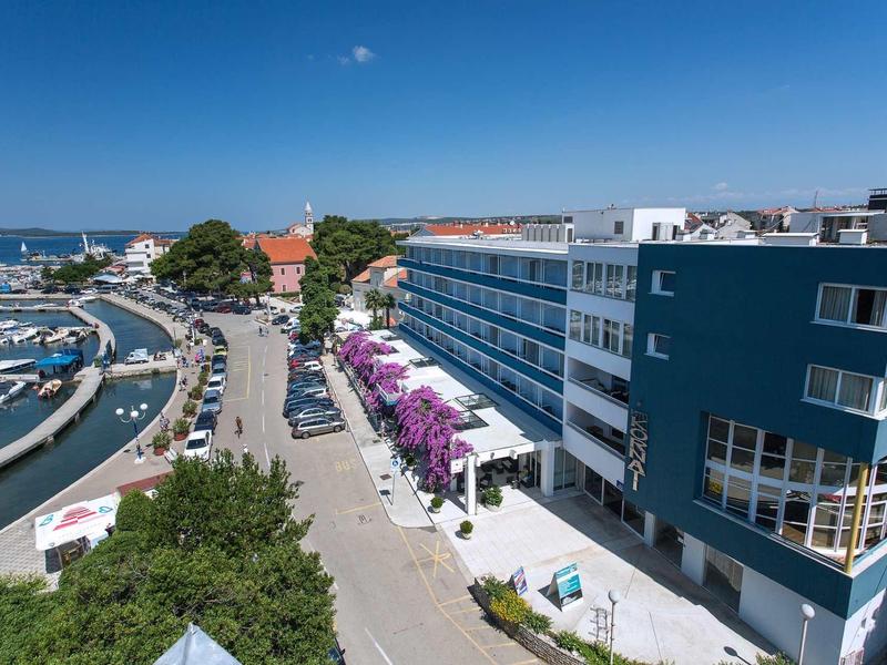 Modern hotelcomplex naast een jachthaven en een straat met bomen en auto's onder een heldere hemel.