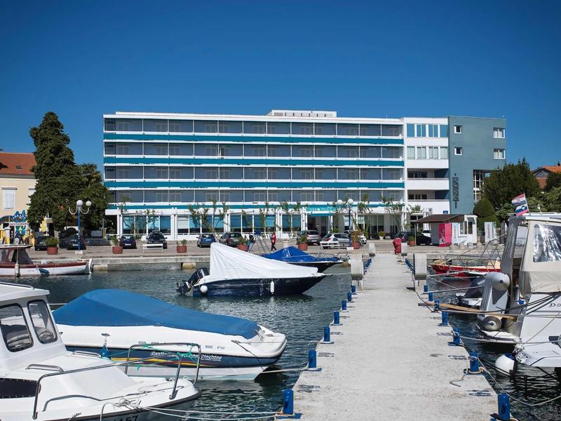 Modern hotelcomplex bij het water met boten aan een steiger onder een heldere lucht.