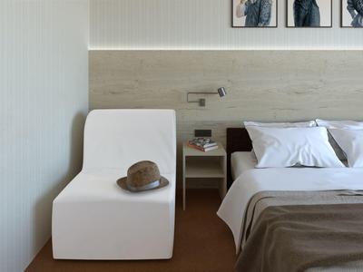 Modern hotelkamer met witte stoel, hoed, nachtkastje en bed met witte kussens.