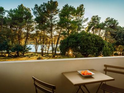 Balkon mit kleinem Tisch und Stühlen, Blick auf Bäume und Meer im Hintergrund bei Sonnenuntergang.