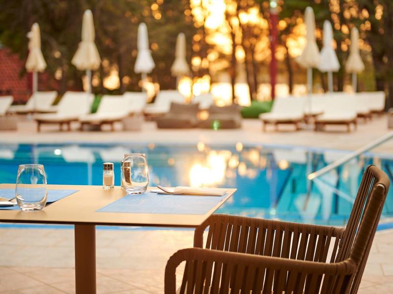 Paisaje de piscina al atardecer con mesa puesta y silla en primer plano bajo sombrillas.