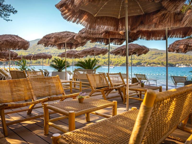 Bar en la playa con sombrillas de paja y sillas de madera junto al agua en un día soleado.