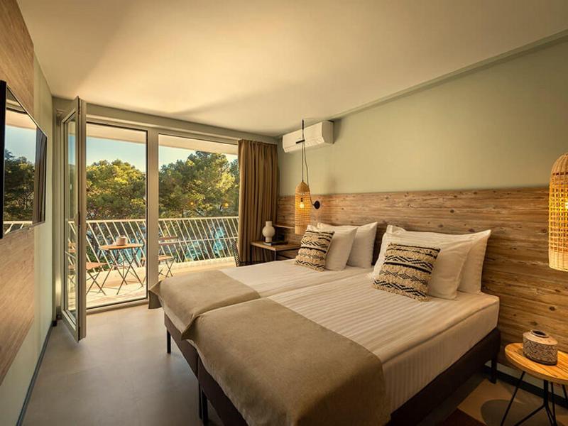 Habitación de hotel moderna con dos camas, detalles en madera y balcón con vista a árboles.