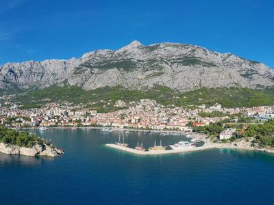 Küstenstadt mit Hafen, umgeben von grünen Hügeln und Bergen, klarer blauer Himmel und ruhiges Meer.