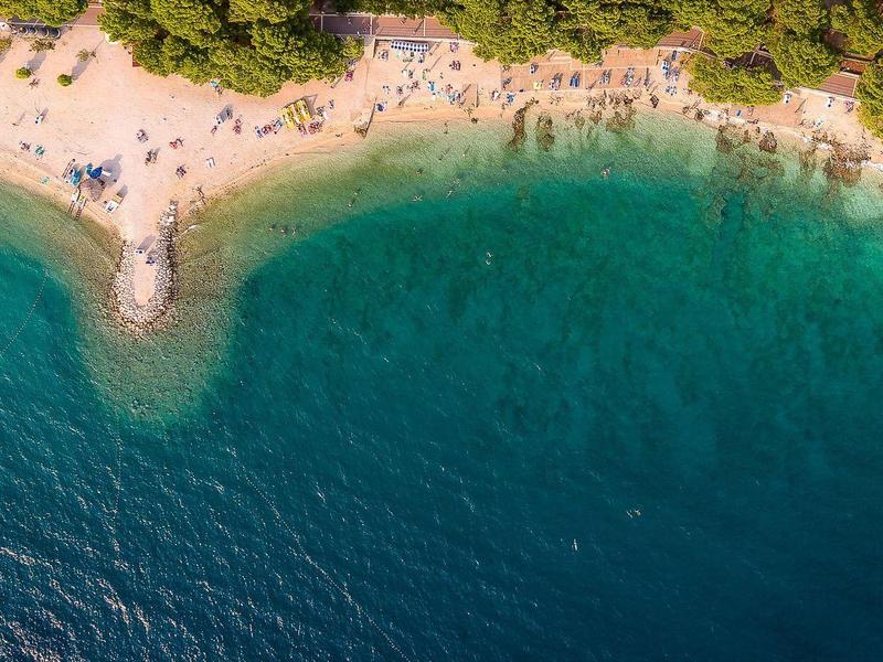Luftaufnahme eines Strandes mit klarem blauem Wasser, Sand und schattenspendenden Bäumen.