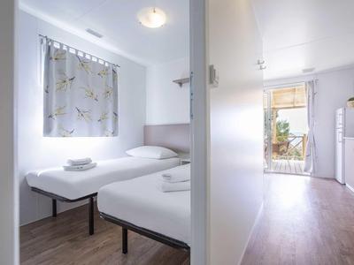 Schlichter, heller Schlafraum mit zwei Einzelbetten, Fenster mit Vorhang und Blick in einen Flur mit Terrasse.