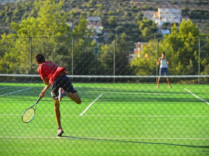 Ein Tennisspieler in rotem Shirt schlägt Ball auf grünem Platz, Gegnerin steht im Hintergrund.