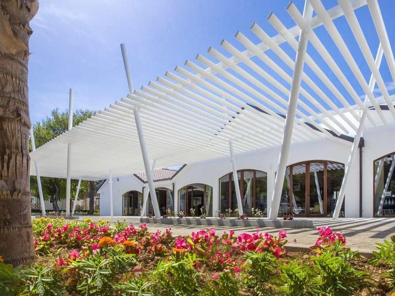 Moderner Pavillon mit weißem Dach und großen Fenstern, davor bunte Blumen und Palmen.