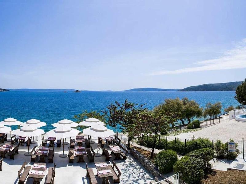 Panorama eines sonnigen Hotelpools mit weißen Sonnenschirmen und Liegestühlen, Blick auf Meer und Inseln.