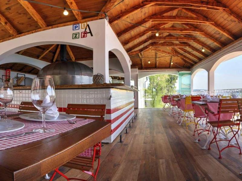 Offene Restaurantterrasse mit Holzdach, Holzboden, karierten Tischtüchern und rotem Stühlen.