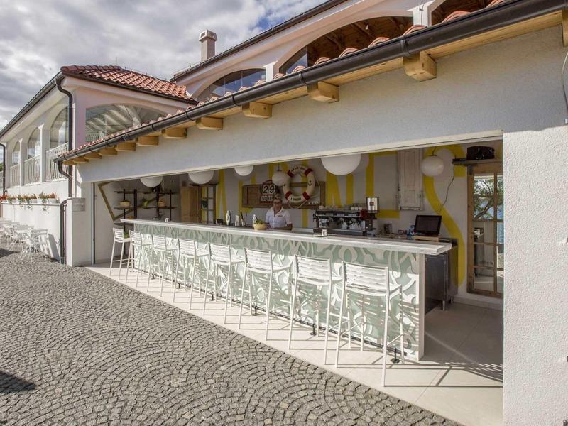 Offener Außenbarbereich mit weißen Barhockern und Ziegelpflasterweg bei bewölktem Himmel.