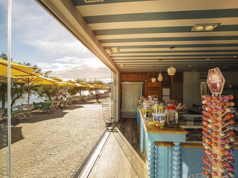 Strandbar mit Snacks, Getränken, Sonnenschirmen und Tischen unter blauem Himmel am Sandstrand.