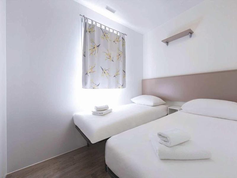 Kleines Zimmer mit zwei Einzelbetten, weißen Wänden, Holzboden und einem Fenster mit Vorhang.