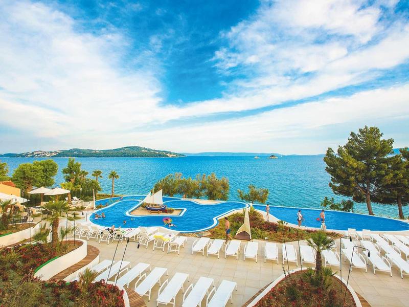 Schwimmbad mit Liegestühlen, Bäumen und Blick auf das blaue Meer unter bewölktem Himmel.