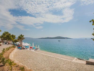 Strand mit Kiesel, Palmen und Blick auf ruhiges blaues Meer unter bewölktem Himmel.