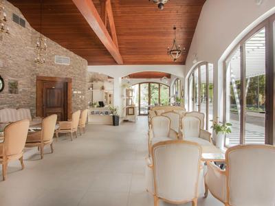 Großer, heller Raum mit Steinwand, Holzbalkendecke und weißen Sitzmöbeln neben großen Fensterbögen.