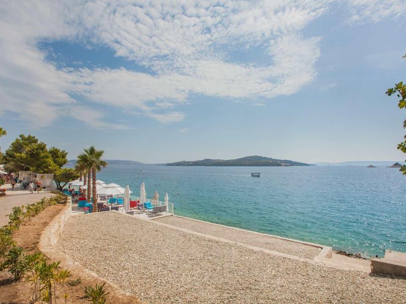 Strand mit Kiesel, Palmen und Blick auf ruhiges blaues Meer unter bewölktem Himmel.