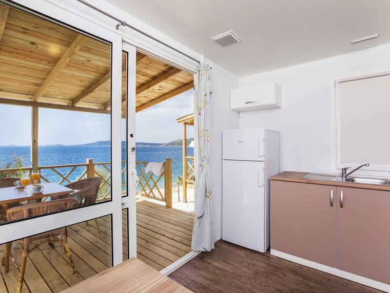 Helle Küche mit kleinem Spülbecken, Kühlschrank und Blick auf überdachte Terrasse mit Meerblick.