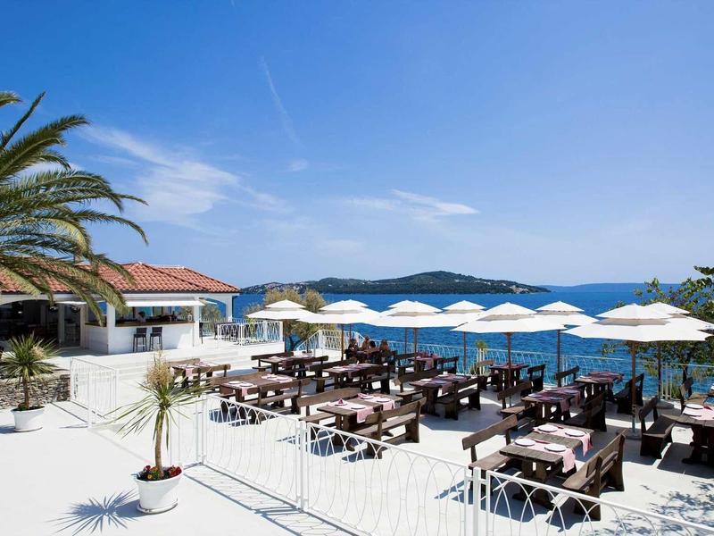 Terrassenrestaurant am Meer mit weißen Sonnenschirmen, Holztischen und Palmen bei klarem Himmel.