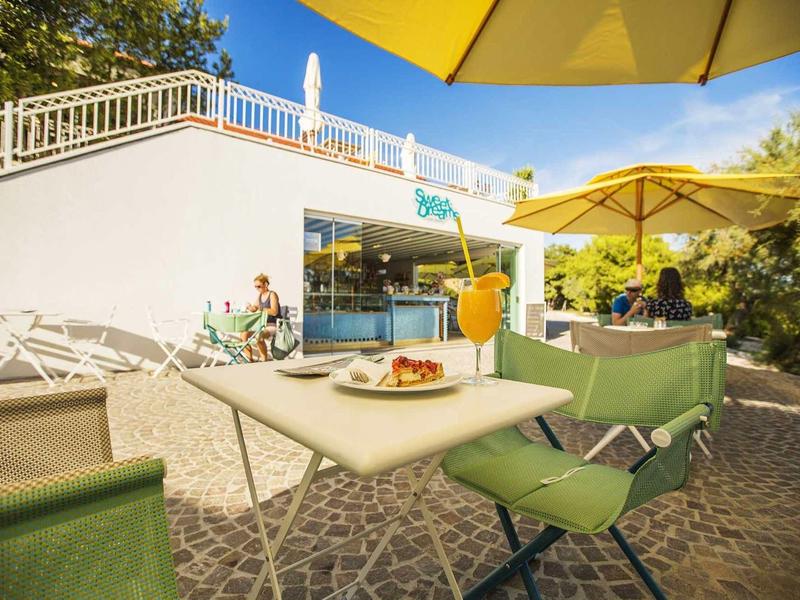 Sonnige Terrasse mit grünen Sesseln, gelbem Sonnenschirm und Blick auf ein modernes Hotelgebäude.