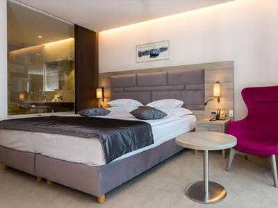 Modernes Hotelzimmer mit großem Bett, grauer Bettdecke, pinkem Sessel und offenem Badezimmer.