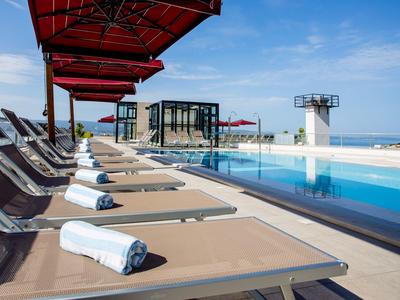 Mehrere Liegestühle mit aufgerollten Handtüchern neben einem Pool auf einer Dachterrasse mit Meerblick.