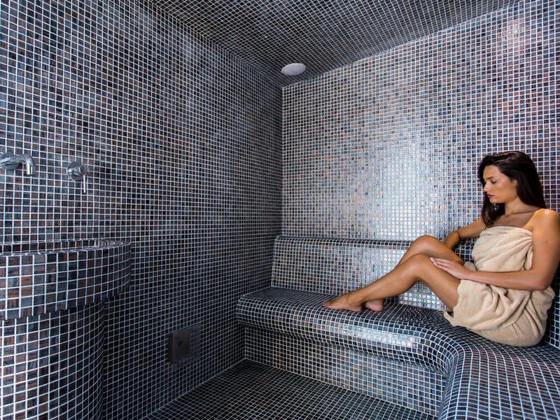 Frau in Spa mit Mosaikfliesen auf Sitzbank und Wänden, entspannt und in Handtuch gewickelt