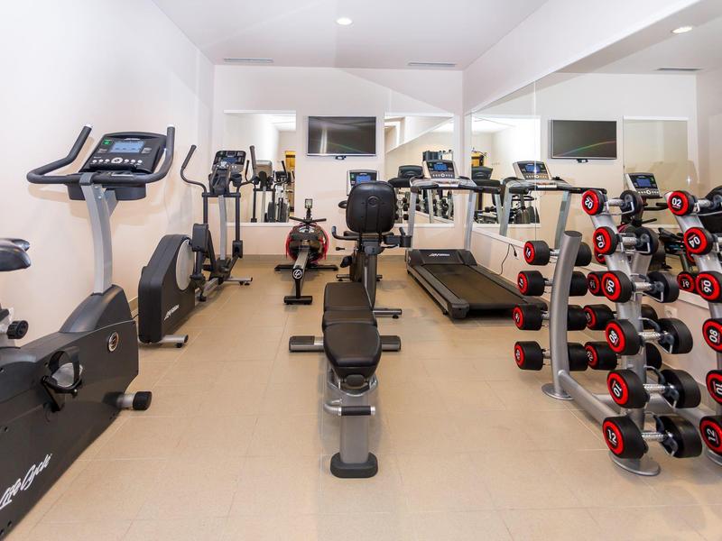 Moderne Fitnessraum mit Trainingsrädern, Laufband und Hanteln vor großen Spiegeln.