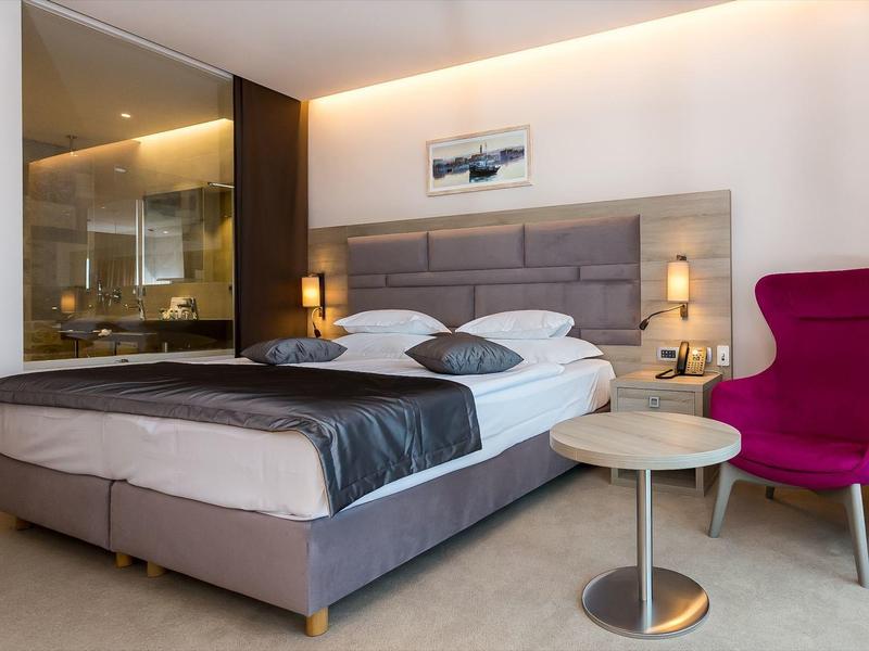 Modernes Hotelzimmer mit großem Bett, grauer Bettdecke, pinkem Sessel und offenem Badezimmer.