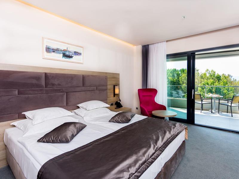 Modernes Hotelzimmer mit großem Doppelbett, rotem Sessel und Balkon mit Grünblick.