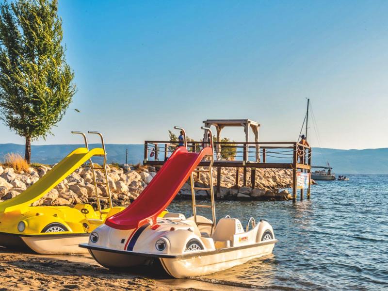 Zwei Tretboote mit Rutschen am Ufer eines Sees neben einem Steg bei klarem Himmel.