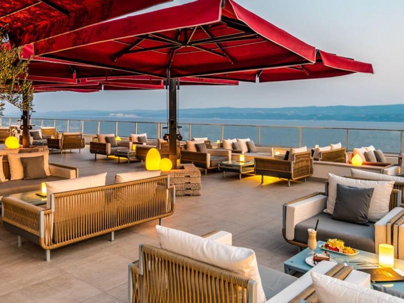Offene Dachterrasse mit bequemen Sitzgruppen, roten Sonnenschirmen und Meerblick bei Abendlicht.