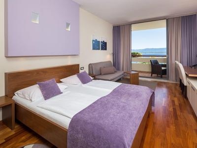 Camera d'hotel con letto matrimoniale, accenti viola, divano e balcone con vista mare