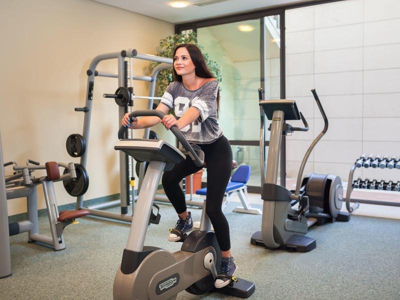 Donna che si allena su una cyclette in una palestra ben illuminata con pesi e attrezzi.