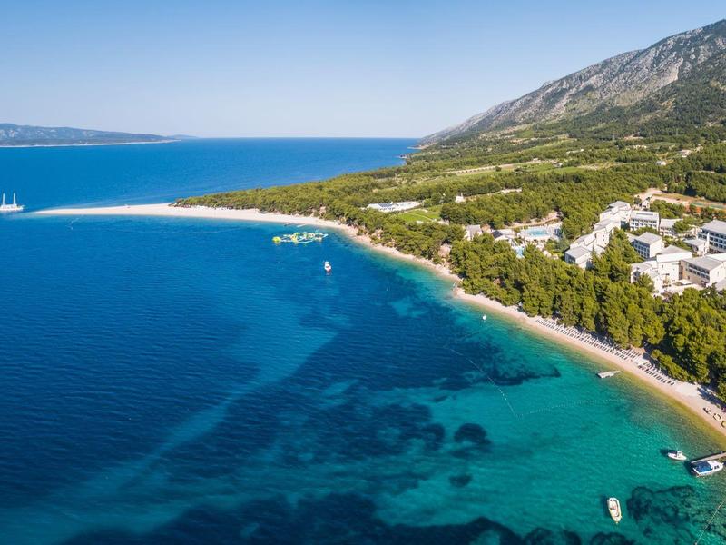 Hotel costiero con spiaggia e acqua blu chiara, circondato da montagne e foreste.