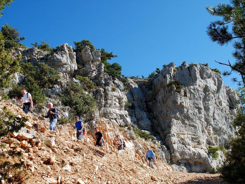 Escursionisti salgono una collina rocciosa accanto a una alta scogliera sotto un cielo blu sereno.