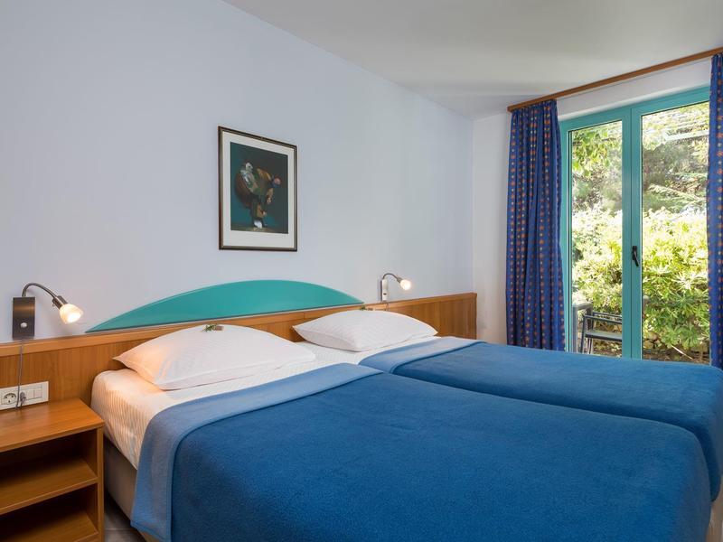 Camera doppia con copriletti blu, vista giardino attraverso grandi finestre, arredamento semplice.