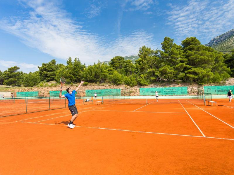 Due persone giocano a tennis su un campo in terra rossa all'aperto circondato da alberi e montagne.
