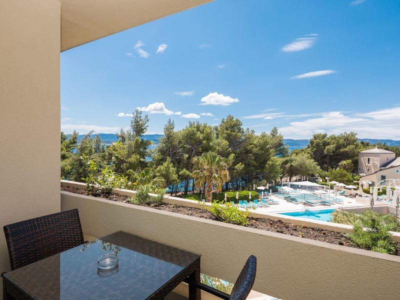 Balcone con tavolo e sedie con vista su piscina, alberi e mare sotto un cielo azzurro.