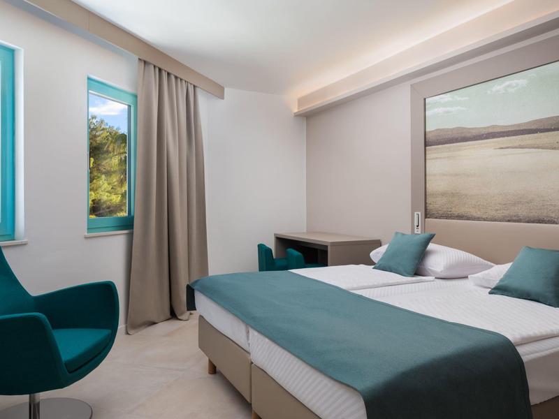 Camera d'hotel moderna con letto matrimoniale, due poltrone blu e finestre con tende.