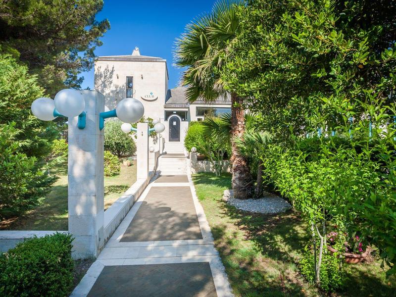 Un sentiero di giardino soleggiato con lampade bianche conduce a un edificio bianco, circondato da piante verdi.