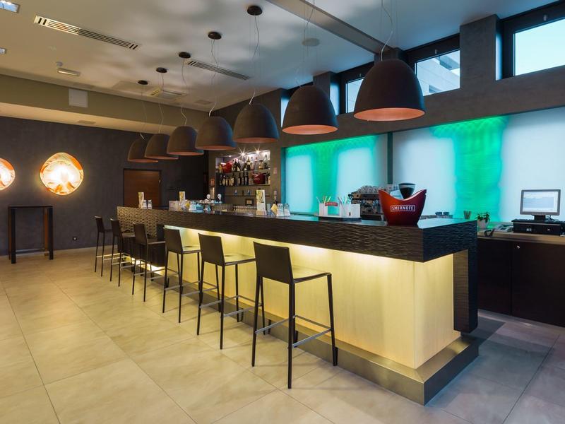 Area bar moderna in hotel con lunghi banconi e sgabelli, illuminata da lampade nere calde.