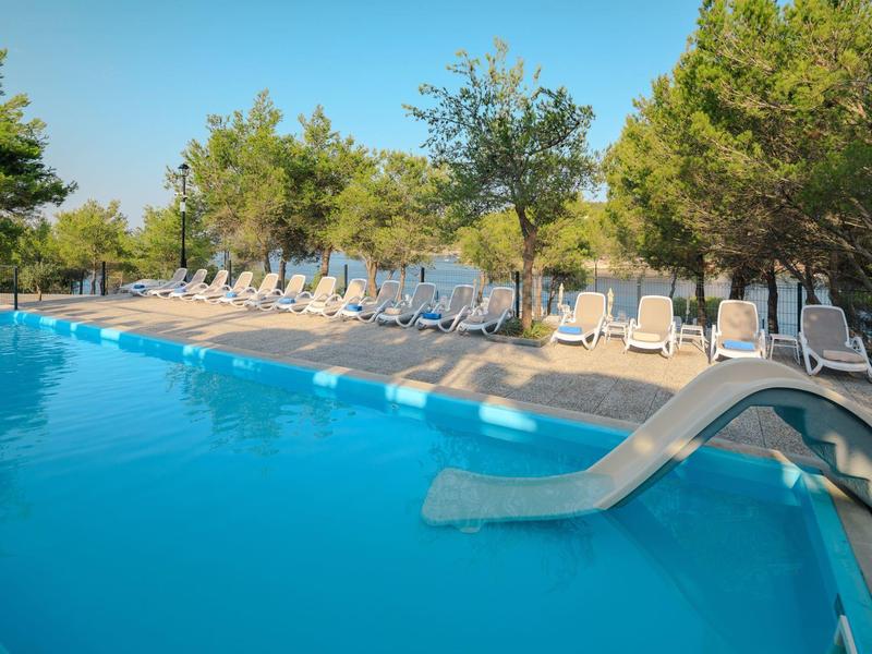 Bazén s modrou skluzavkou a lehátky pod stromy za slunečného dne.