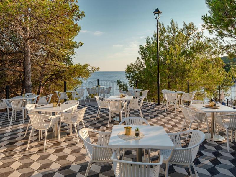 Terasa s bílými stoly a židlemi obklopená stromy s výhledem na moře.