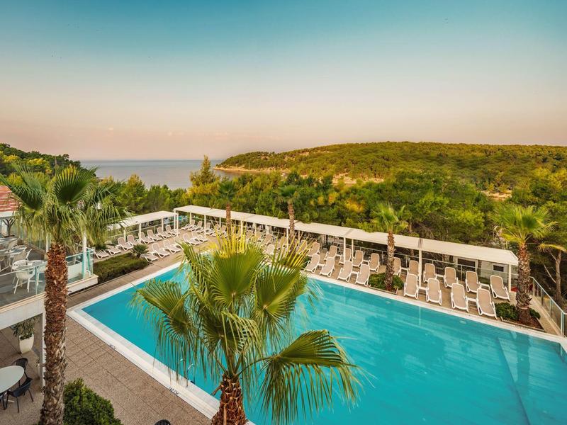 Pohled na obdélníkový bazén s bílými lehátky a zelenou krajinou v pozadí.