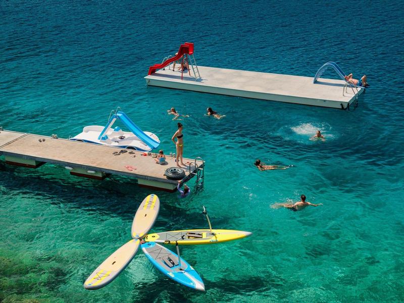 Persone che nuotano in acqua turchese vicino a due pontili con scivolo e kayak.