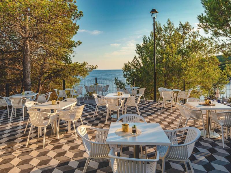 Terrazza dell'hotel con tavoli e sedie bianchi sotto pini, vista mare.